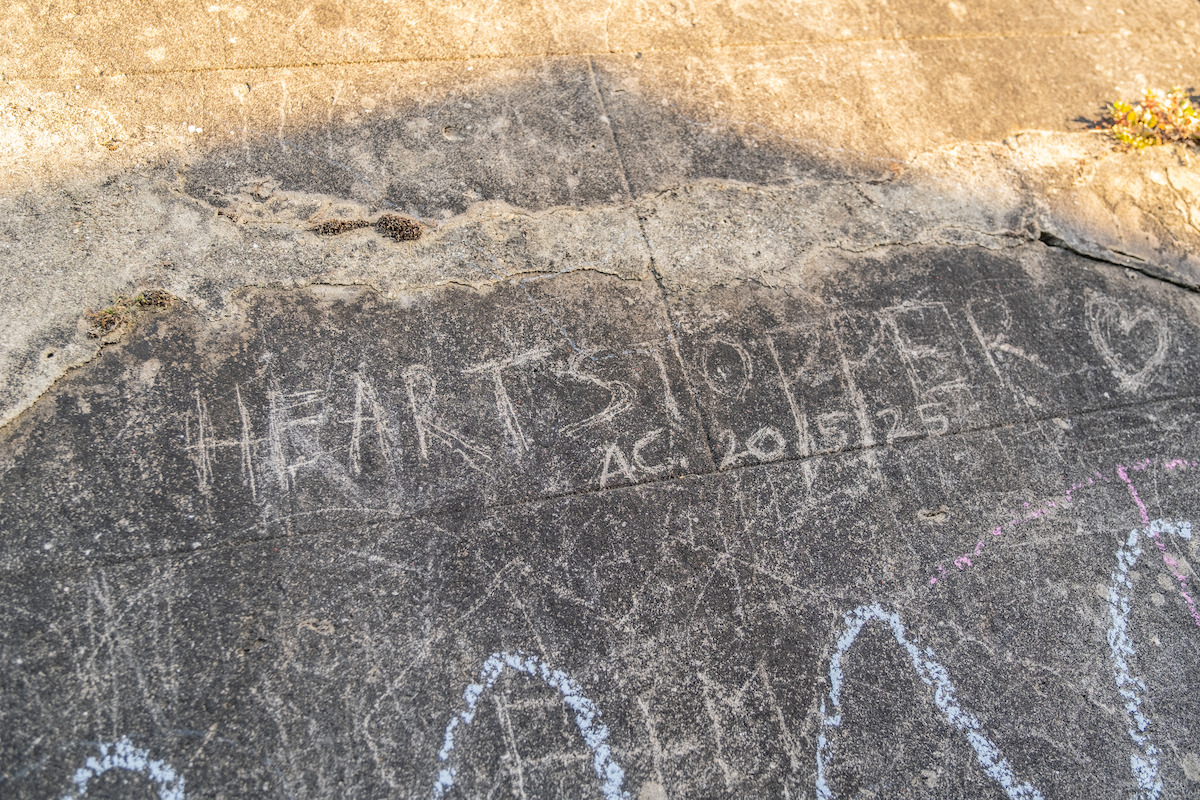 The image shows the word "HEARTSTOPPER" and a heart drawn with chalk on a worn concrete sidewalk, with additional faded chalk doodles and plants visible along the edge. Outdoor and sunny environment.