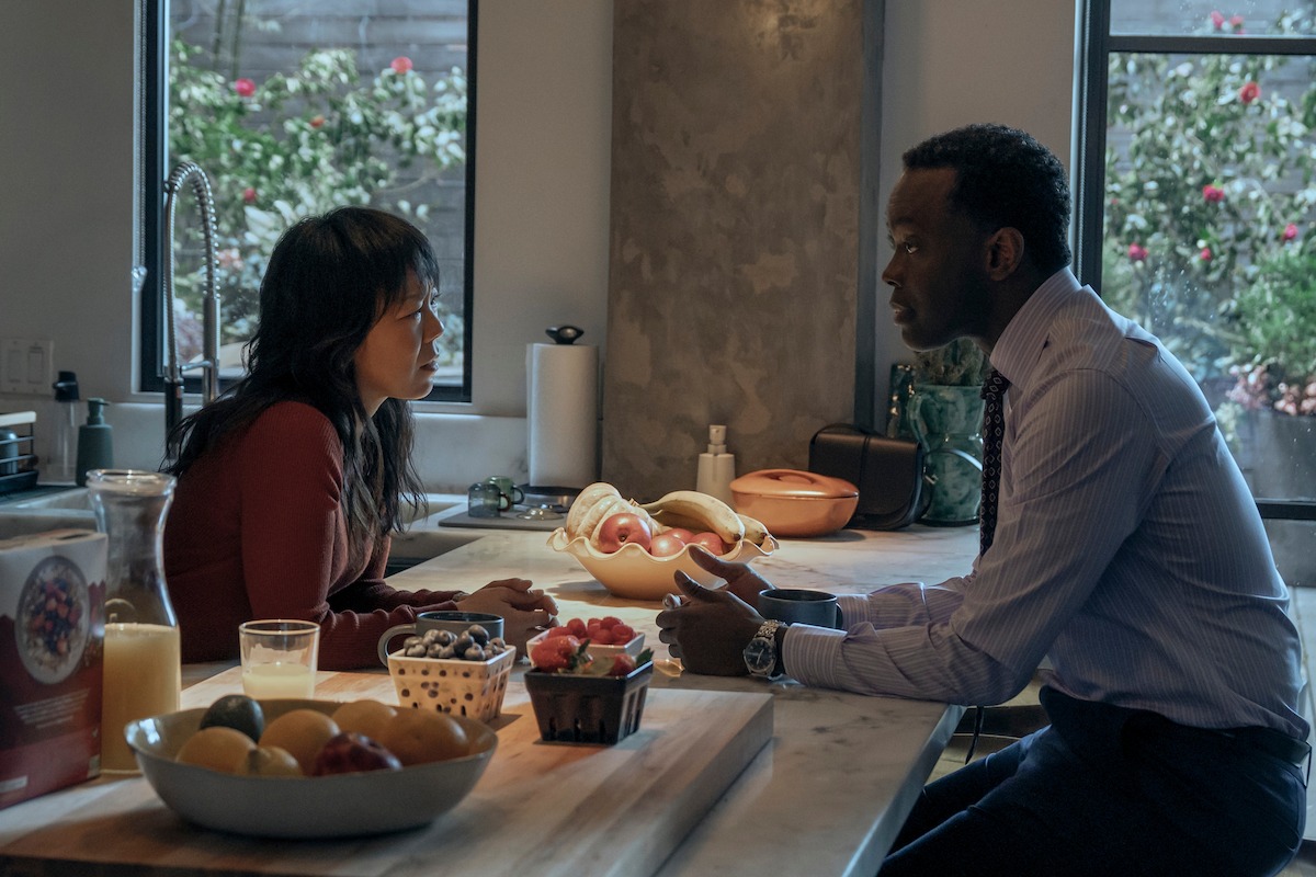 Two people sitting at a kitchen island having a serious conversation, surrounded by fruit, juice, and soft daylight from large windows with a view of a garden outside.