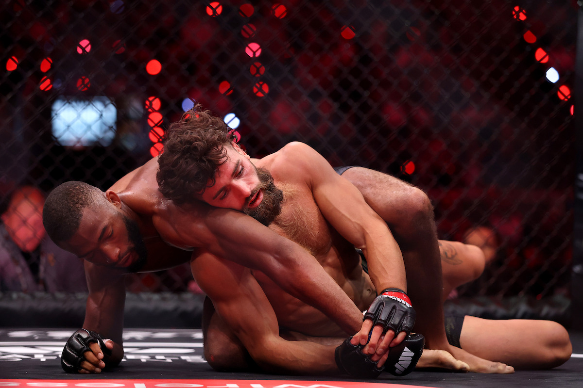 Two mixed martial artists grapple on the mat inside a caged octagon during a professional MMA match, with the crowd and cage visible in the background under arena lighting.