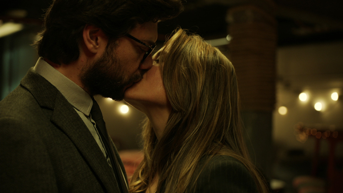 A man and woman in formal attire kiss in a dimly lit indoor setting with warm lights and blurred background, suggesting an intimate or romantic moment in an upscale environment.