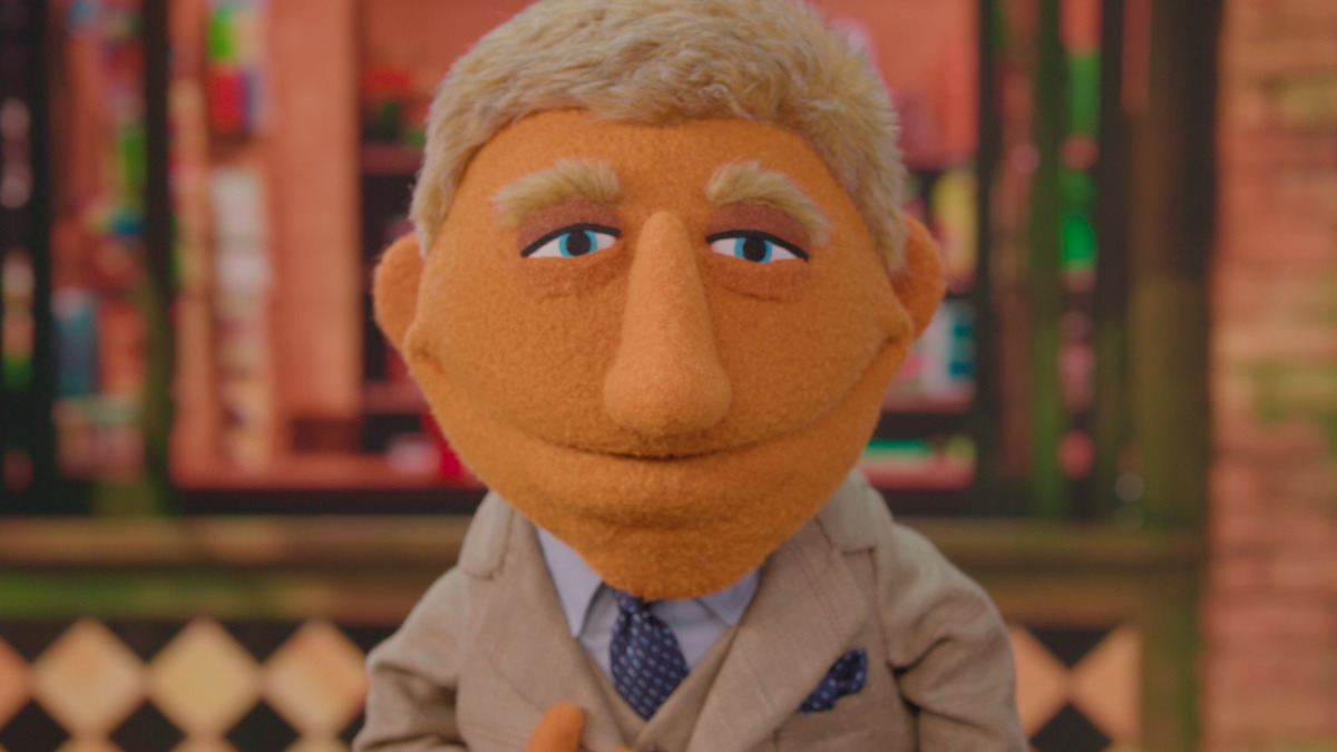 A blond puppet in a beige suit smiles in a warmly lit, indoor setting with a colorful, blurred background, creating a welcoming, cheerful mood.