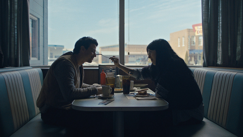 Two people sit in a vintage-style diner booth sharing a meal, with one feeding the other. Daylight streams through large windows, and condiments are on the table, creating a cozy, intimate atmosphere.