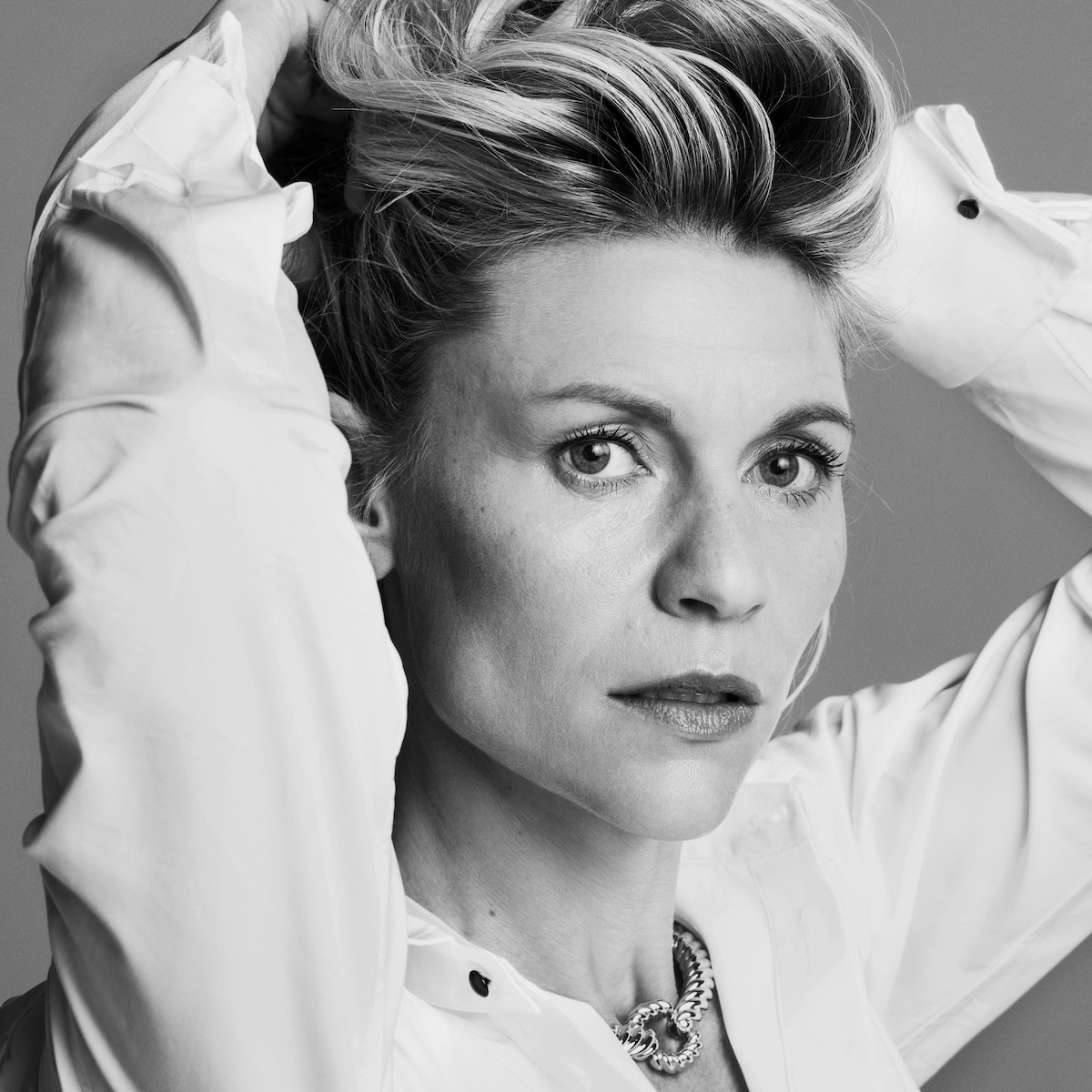 Black and white portrait of a woman in a white shirt, hands in her hair, looking directly at the camera with a serious expression, minimal background, conveying a contemplative and intense mood.