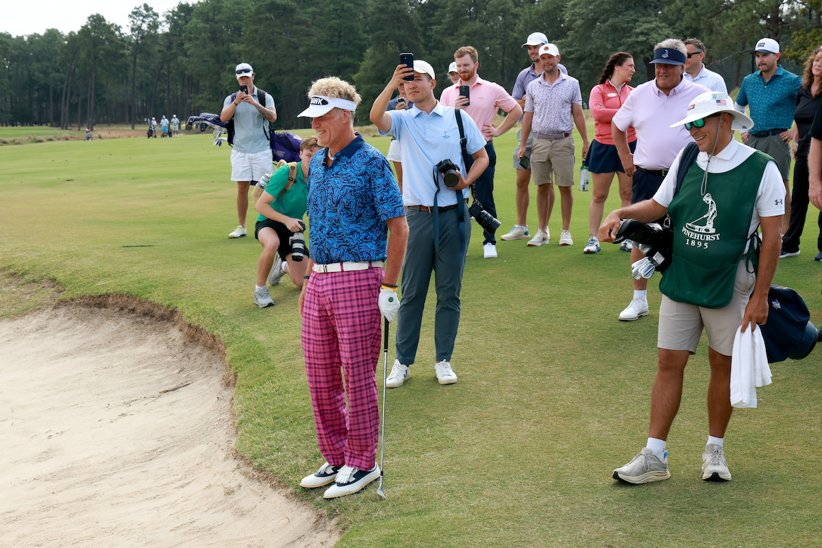 Golfer in colorful outfit stands near a sand bunker on a golf course, holding a club, with caddie beside him and a group of spectators and photographers watching in the background under partly cloudy skies.