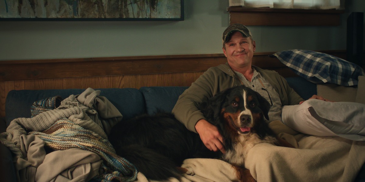 A man wearing a cap sits on a blue couch in a cozy living room, smiling while petting a large Bernese Mountain Dog. The environment is casual with blankets and pillows scattered around.