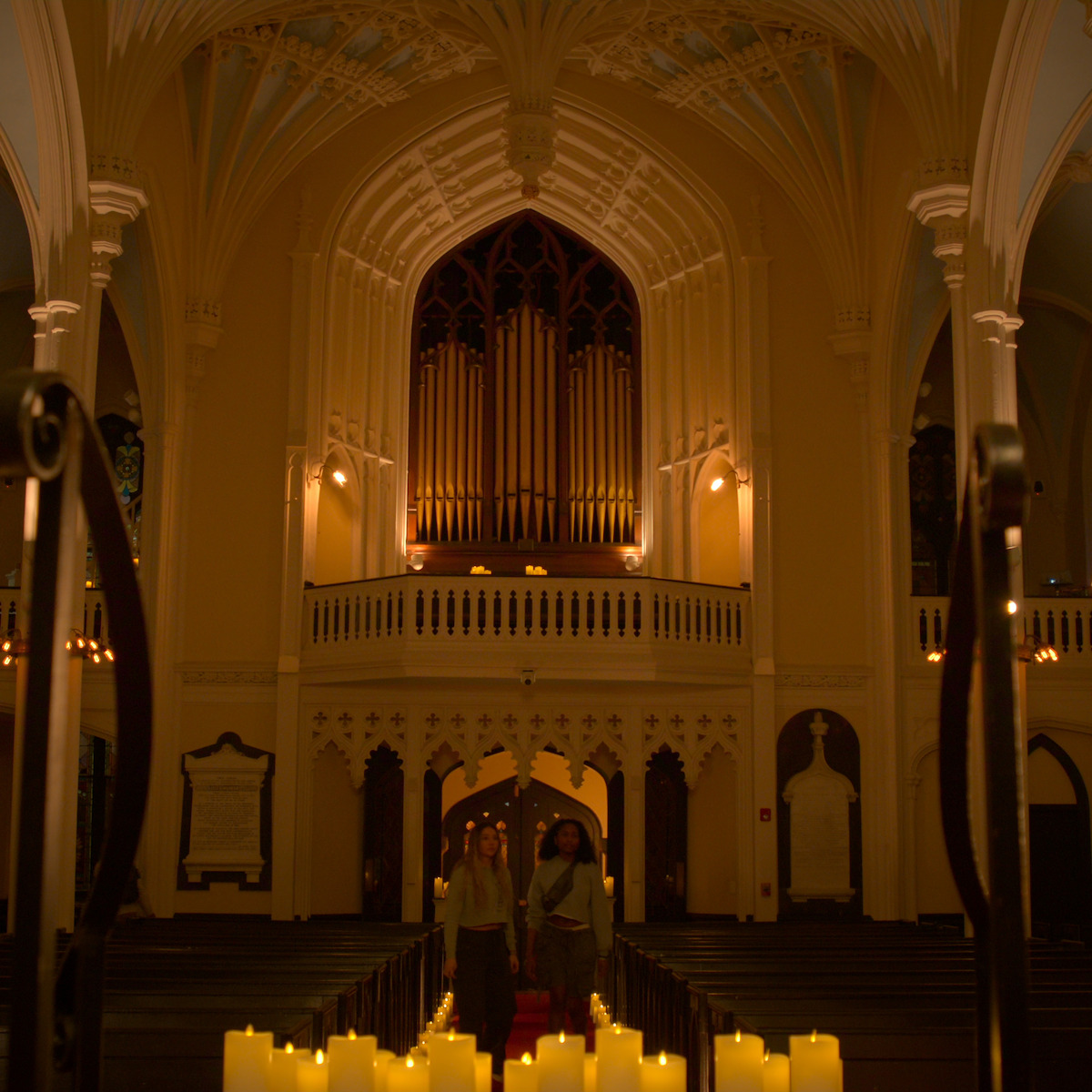 Duas pessoas atravessam o corredor de uma igreja mal iluminada com teto alto e abóbadas, muitas velas iluminando o caminho. O lugar tem arquitetura sofisticada, com colunas, vitrais e um grande órgão de tubos acima da entrada.