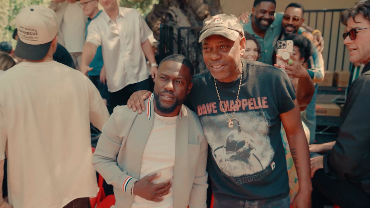 Two men pose for a photo at an outdoor gathering, surrounded by a cheerful crowd. One wears a graphic t-shirt and hat, the other a light jacket. People in the background are smiling, some holding phones, in a sunny, casual setting.