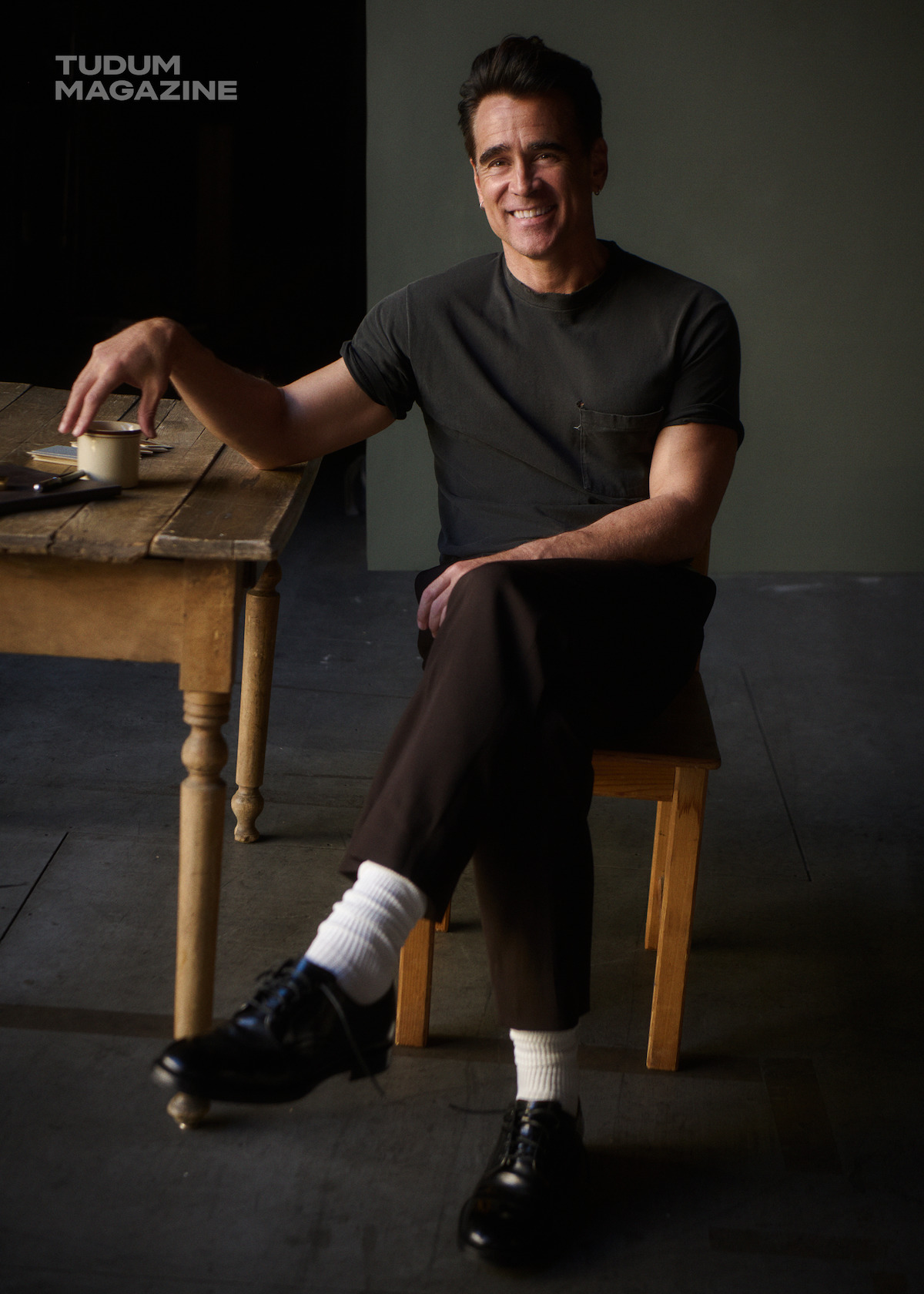 Colin Farrell wears a grey shirt and brown pants with black derby shoes and white socks as he sits at a table with a coffee cup.