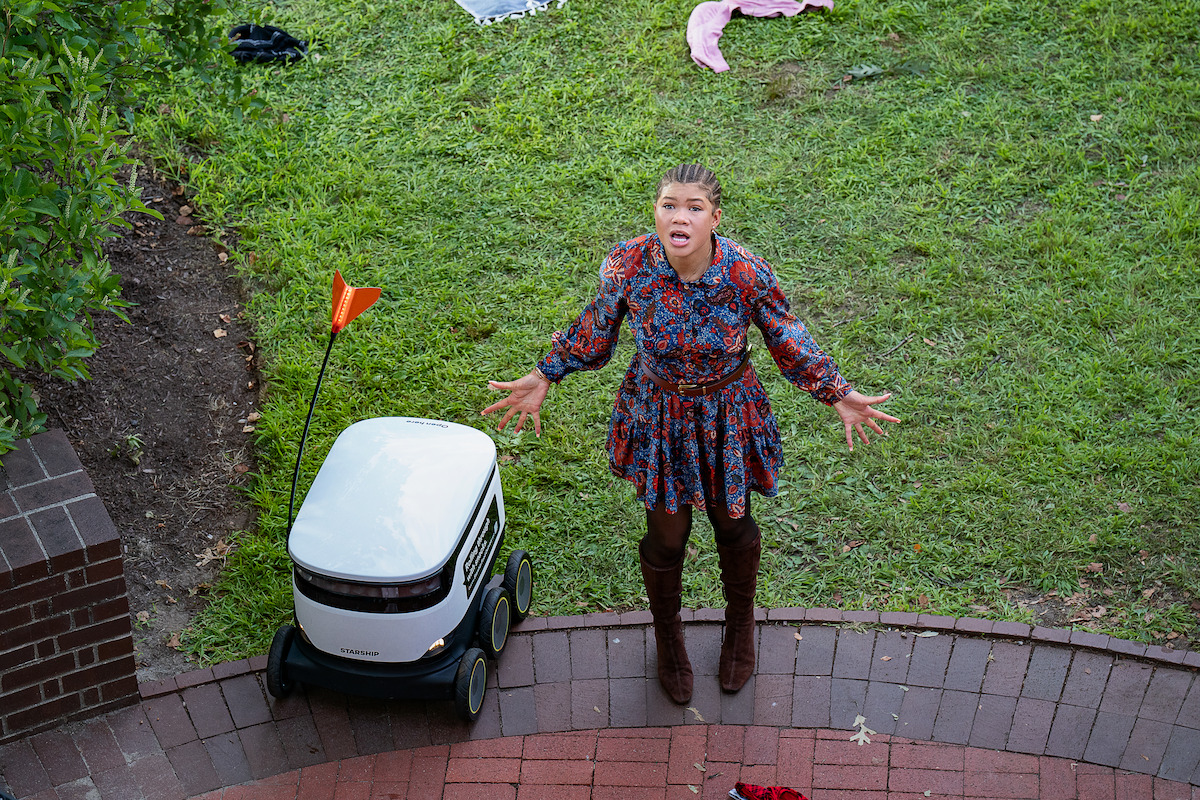 Woman in a floral dress and boots standing on a brick walkway next to a small delivery robot with an orange flag, in a grassy outdoor park setting, looking upward with surprised expression.