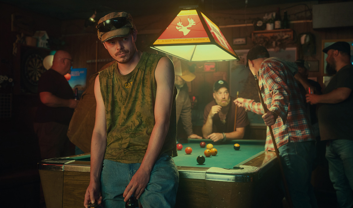 Man in sleeveless shirt and cap leaning on pool table in dimly lit bar, with several people playing pool and socializing in the background under a hanging lamp.