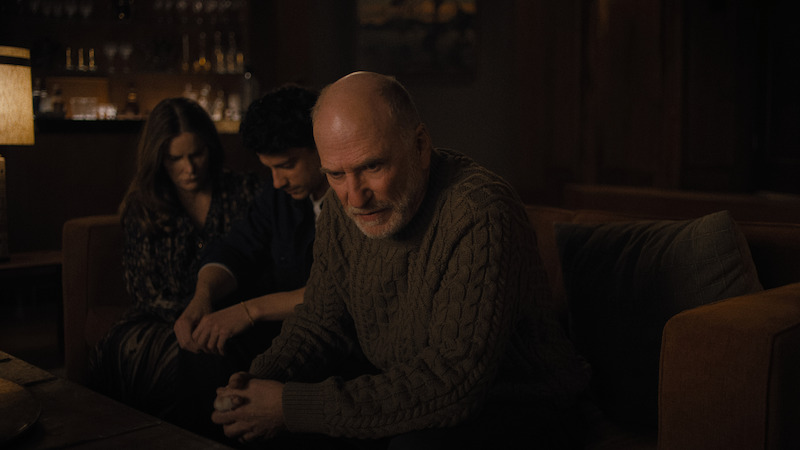 Three people sit on a dimly lit couch, looking serious and contemplative in a cozy living room with warm lighting and shelves with glassware in the background.