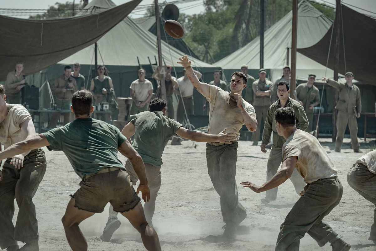 Soldiers in vintage uniforms play football on a dusty field surrounded by military tents, with other soldiers watching, suggesting a break in a wartime or military camp setting.