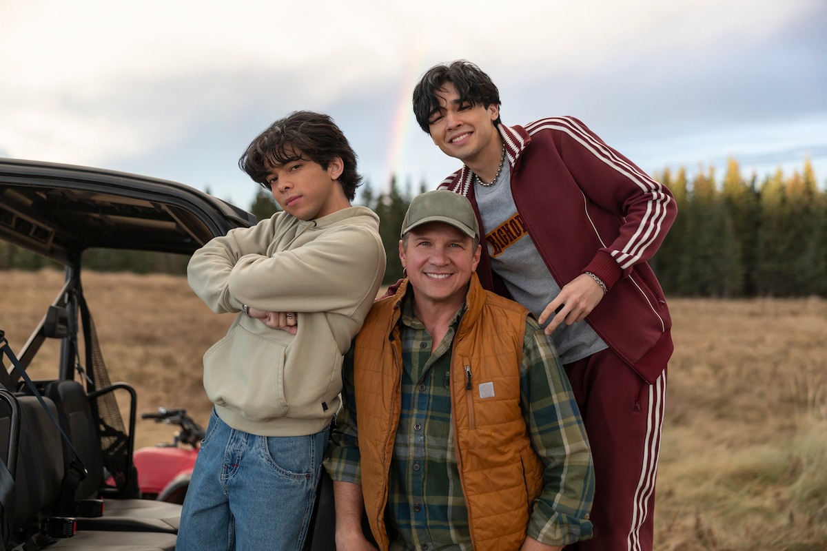 Three people smile outdoors next to an ATV in a grassy field with trees and a rainbow in the background. One person stands with arms crossed, another wears a cap and vest, and the third is in a tracksuit.