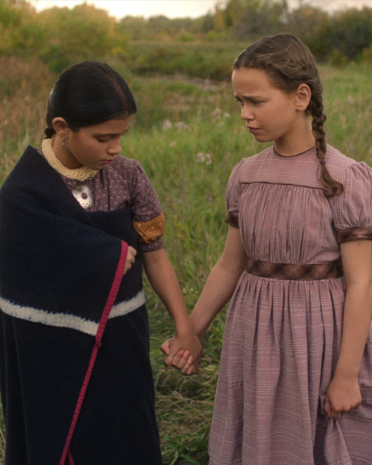 Two young girls in vintage dresses stand in a grassy field holding hands; one wears a dark shawl and looks down, while the other, with braided hair, looks at her with concern. The setting appears rural and peaceful.