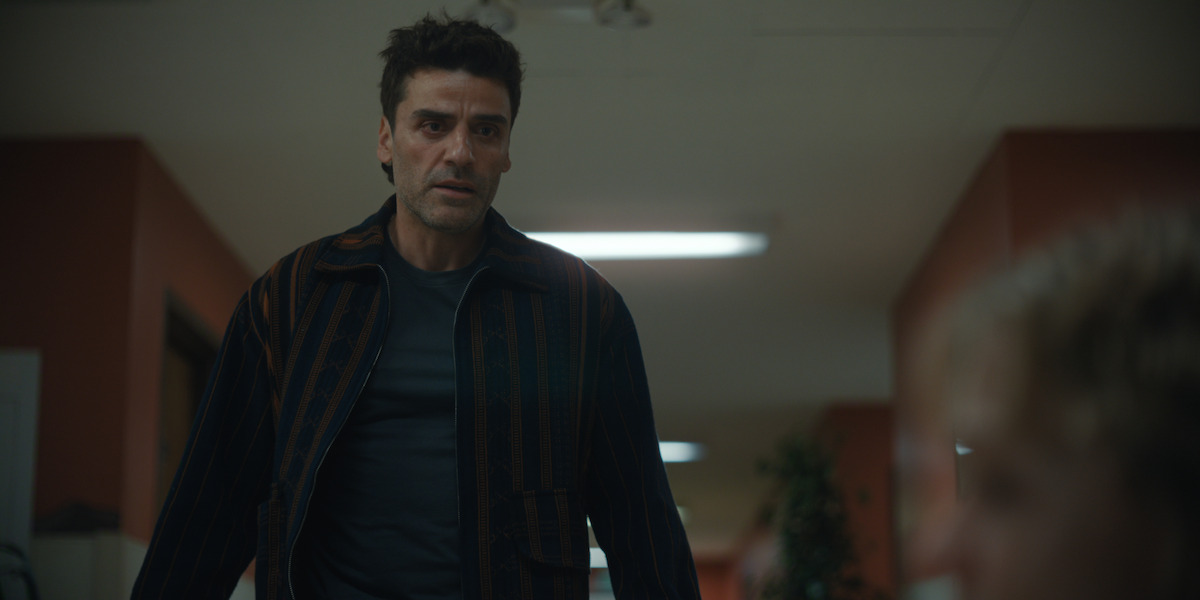 Man with serious expression stands in a dimly lit hallway with reddish walls and fluorescent lights, looking toward a blurred figure in the foreground, evoking tension or concern in an indoor institutional setting.