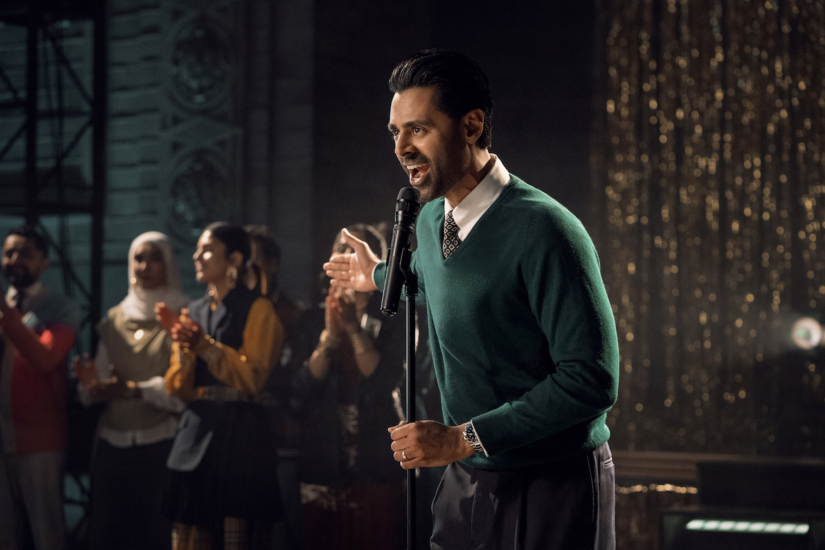 Man in green sweater speaks into microphone on stage, with an audience of people clapping and watching in a warmly lit, festive indoor environment with golden decorations in the background.