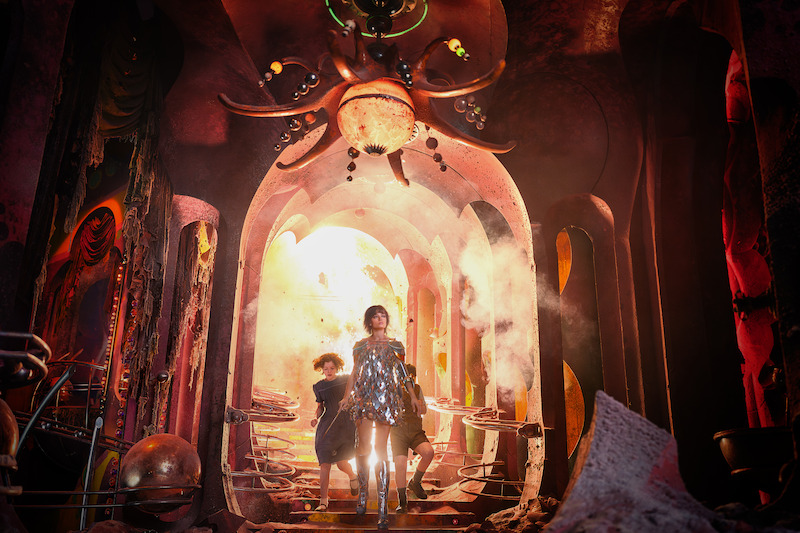 Three people, including a woman in a shiny silver dress, walk through a surreal, sci-fi corridor with dramatic lighting, smoke, and decor in a futuristic or fantastical setting.