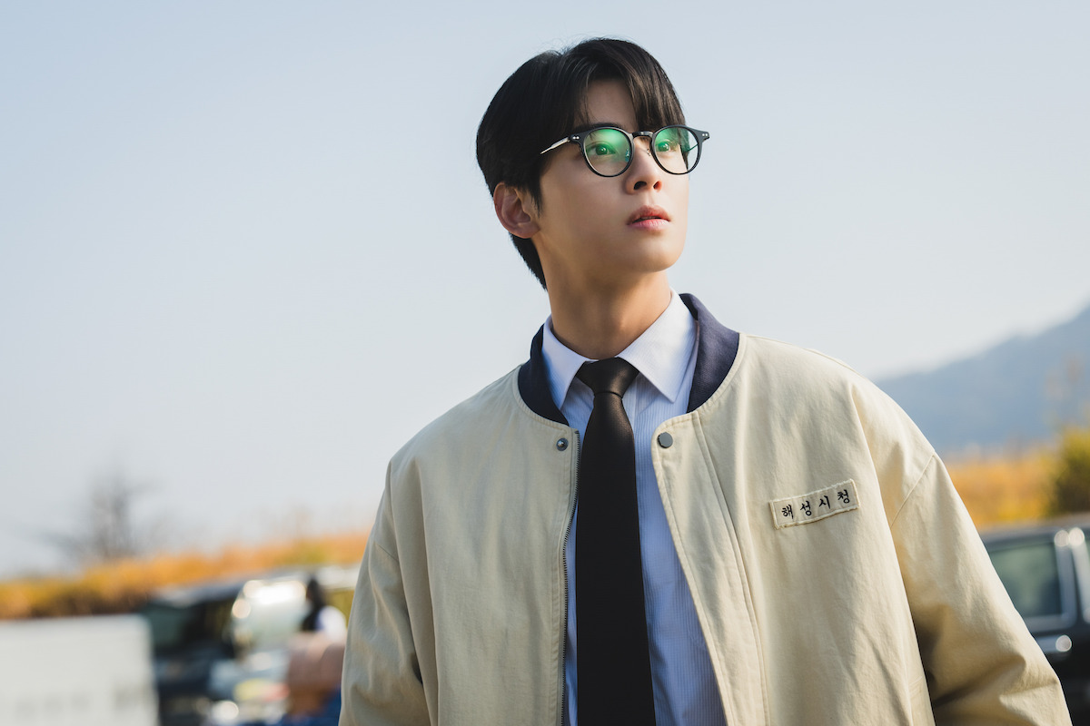 Young man wearing glasses, a tie, and a beige jacket stands outdoors on a clear day, looking into the distance. The background shows blurred cars and autumn foliage with mountains under a blue sky.