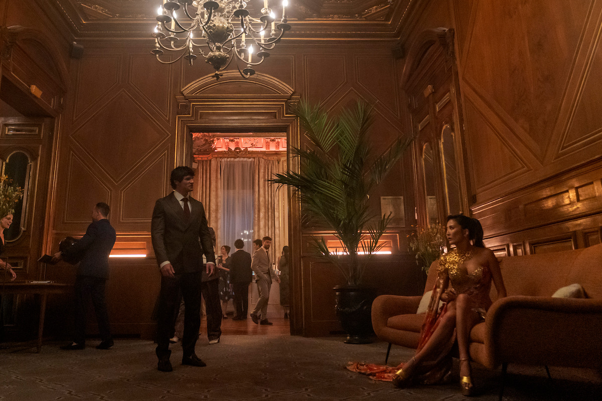 A man in a suit stands in an elegant, wood-paneled lounge with a chandelier, while a woman in a gold dress sits on a couch; other people are visible in the background socializing at a formal event.