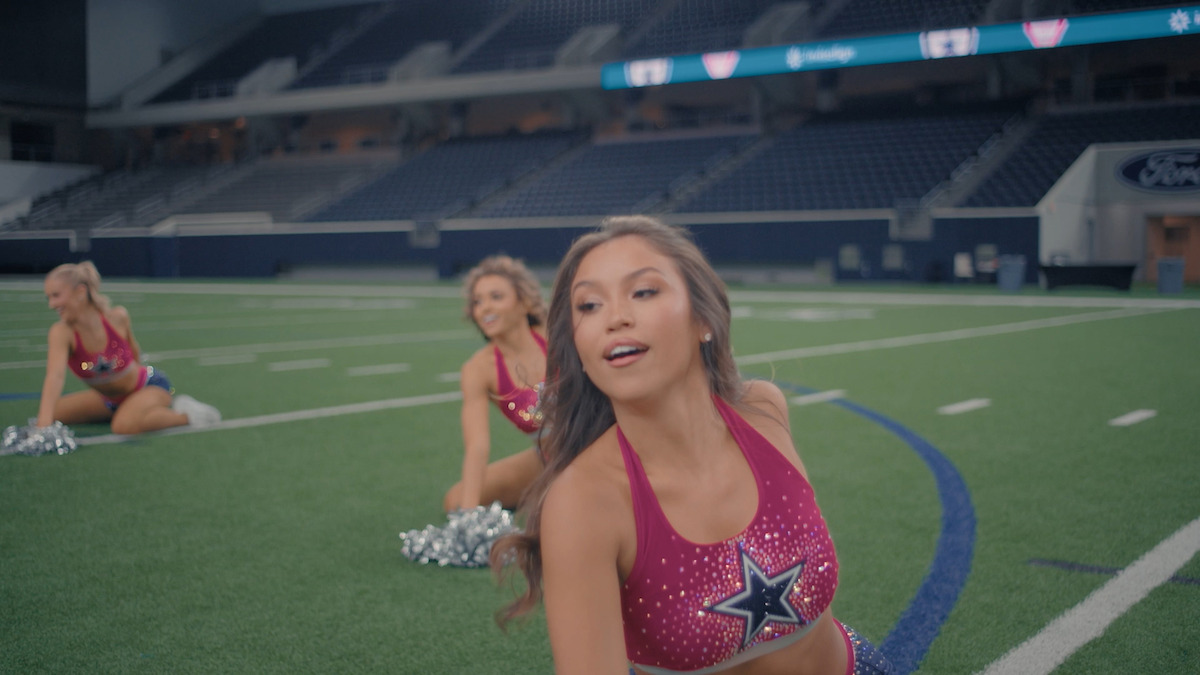 Three cheerleaders in matching pink uniforms perform a routine on a football field inside a large, empty stadium. The focus is on the cheerleader in the foreground, with two others posing in the background.