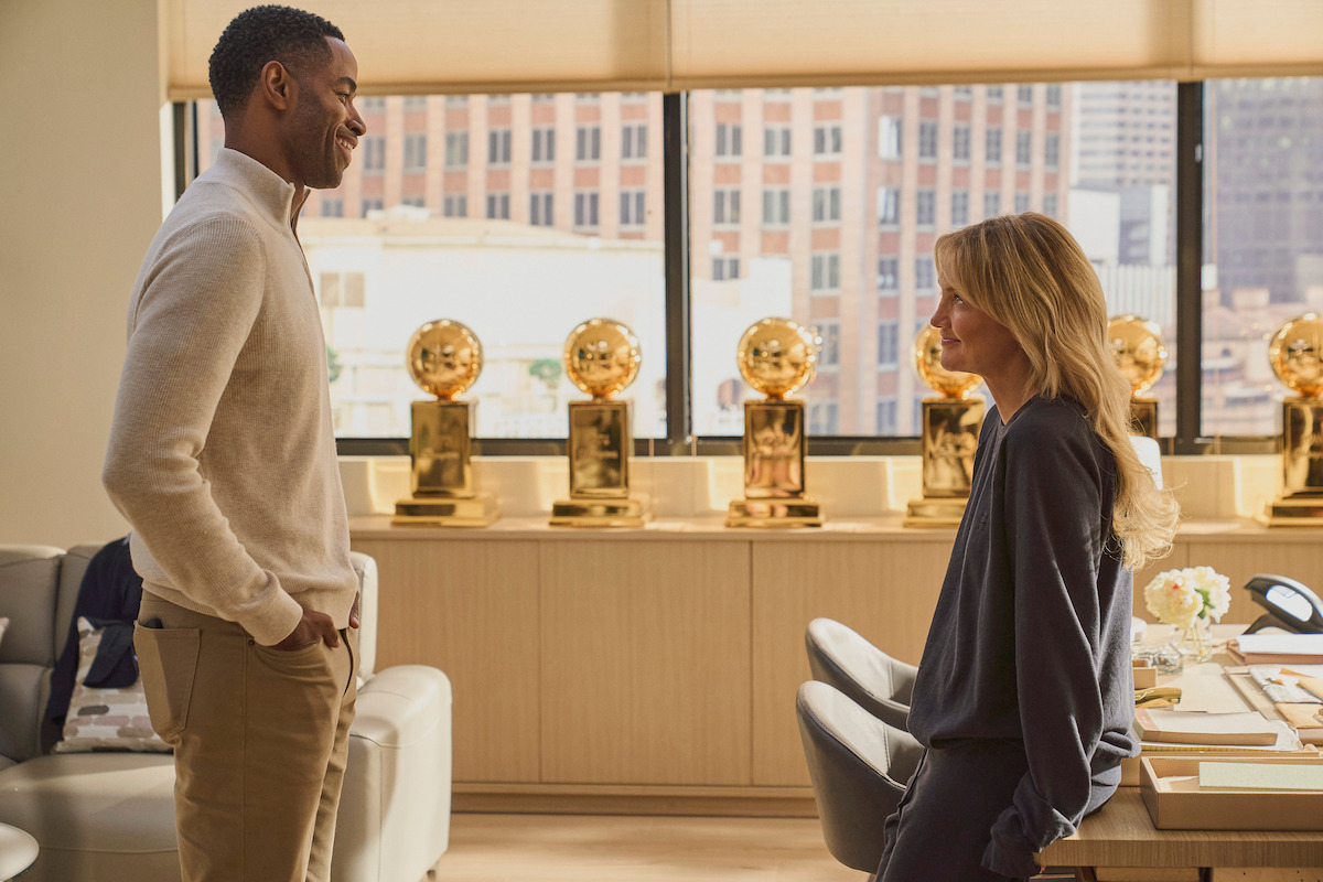 Two people talking in a modern office with large windows and several basketball trophies displayed on a shelf in the background.