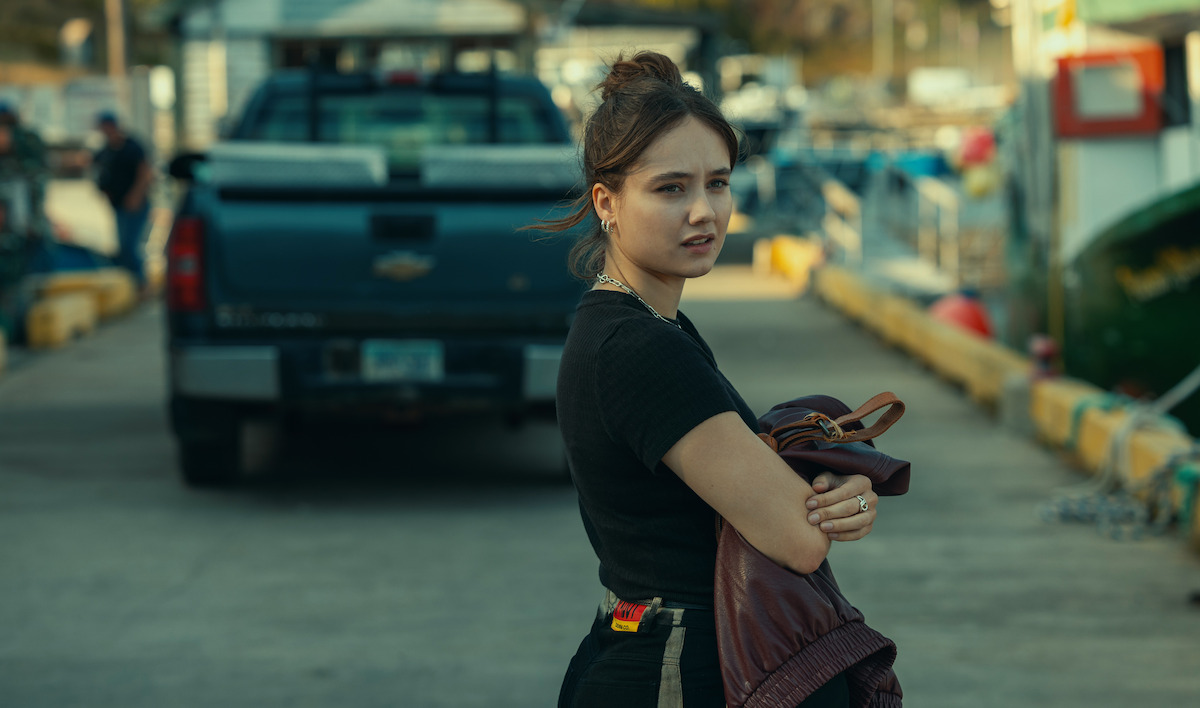 Young woman holding a brown jacket stands on a dock near parked trucks, looking back with a serious expression, in an outdoor marina environment with boats and industrial equipment in the background.