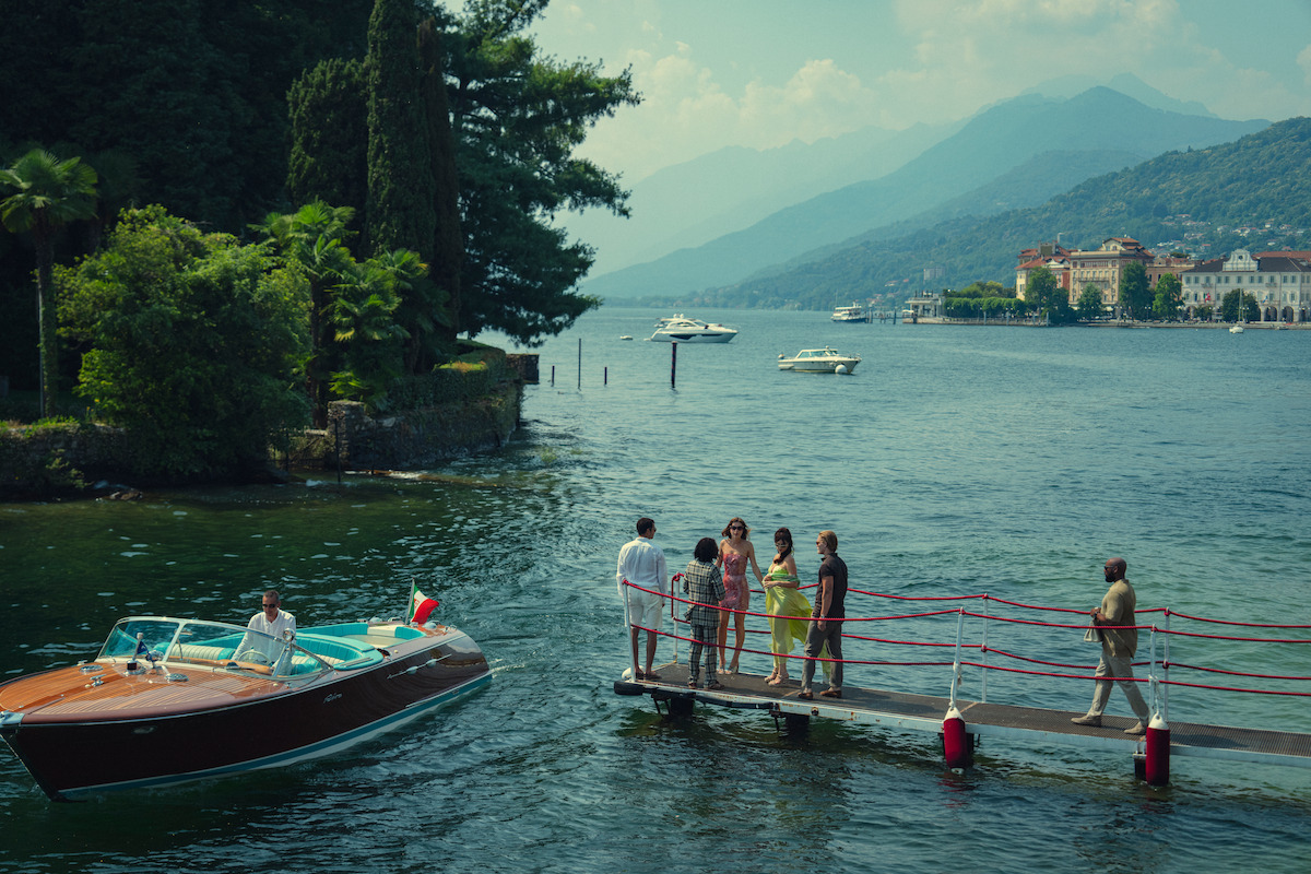 A group of people stand on a dock by a scenic lake as a wooden motorboat approaches. The background features lush greenery, distant boats, mountains, and elegant lakeside buildings under a hazy sky.