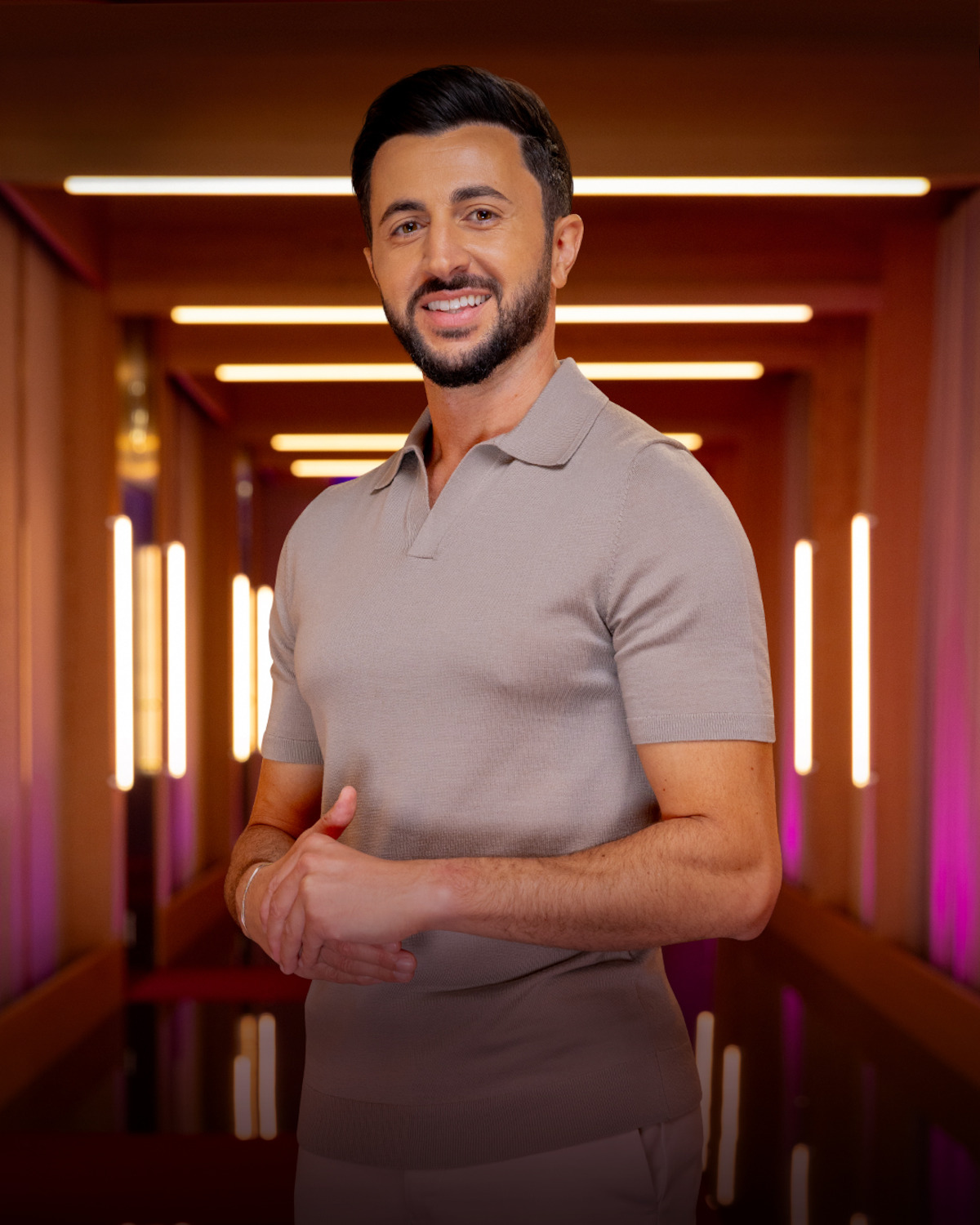 Smiling man with dark hair and beard stands in a warmly lit modern hallway with wooden accents and vertical light fixtures, wearing a short-sleeve beige polo shirt.