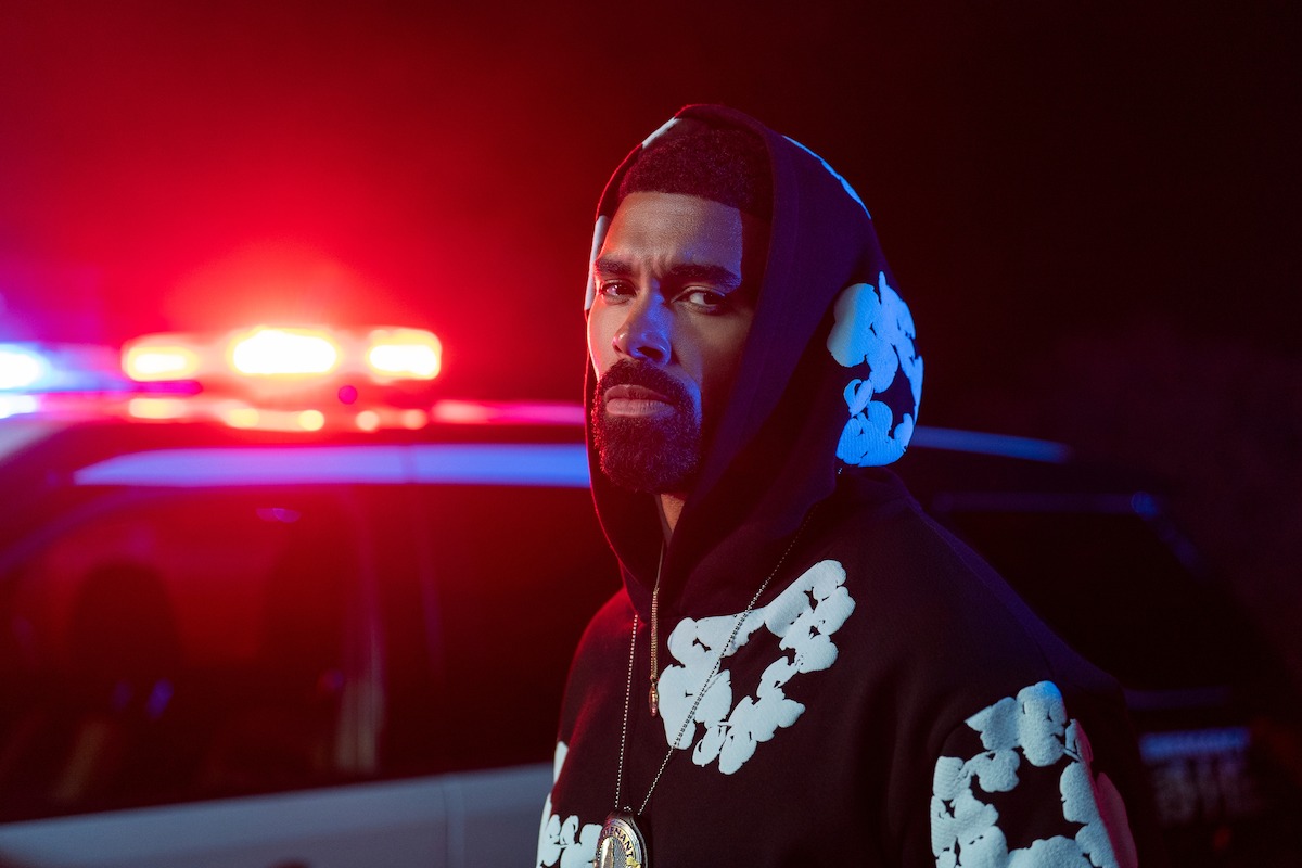 Man in hooded sweatshirt standing in front of police vehicle with flashing red and blue lights at night, serious expression, nighttime outdoor setting.
