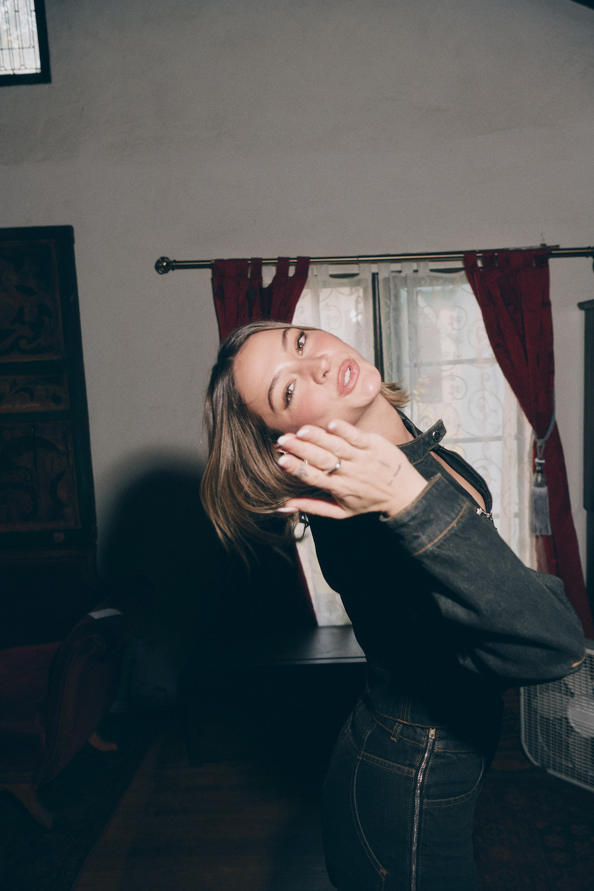 Woman in a dark denim outfit posing playfully indoors, blowing a kiss. The room features red curtains, a window with metal grilles, wooden furniture, and soft, natural lighting.