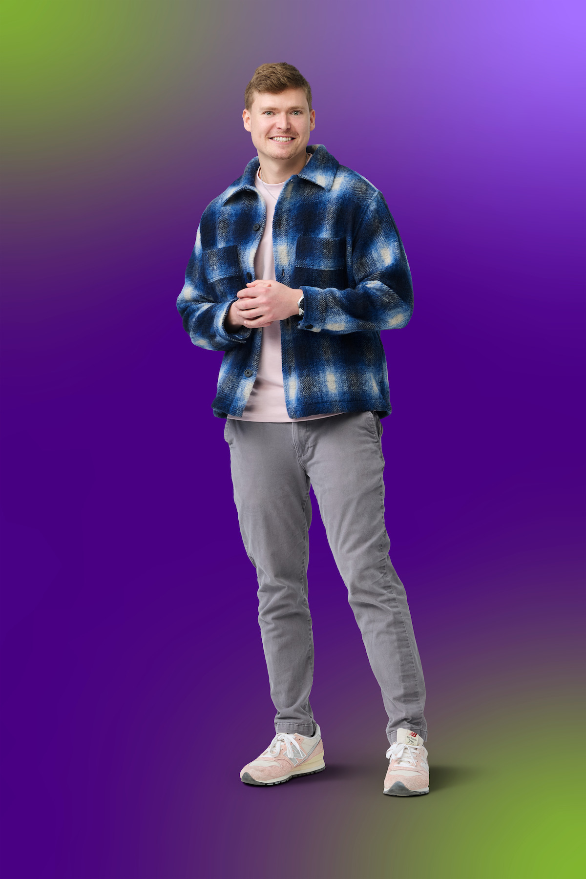 Smiling man wearing a blue plaid jacket, light shirt, gray pants, and sneakers stands against a vibrant purple and green gradient background.