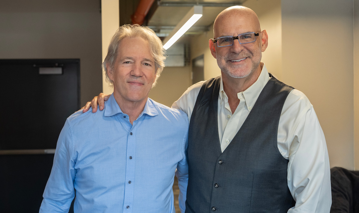 Two smiling men stand side by side in an indoor office hallway, one with a hand on the other's shoulder. Both are dressed in business casual attire, with neutral-colored walls and a door in the background.
