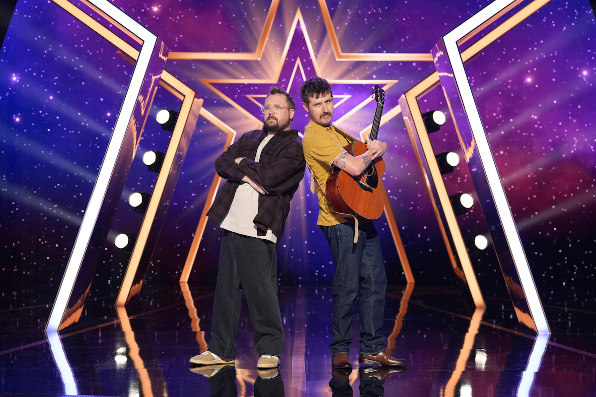 Good Cop and Rad Cop pose back-to-back on a stage with a large star backdrop and stage lights; one holds a guitar, both look confident, under dramatic purple and gold lighting in a talent show environment.