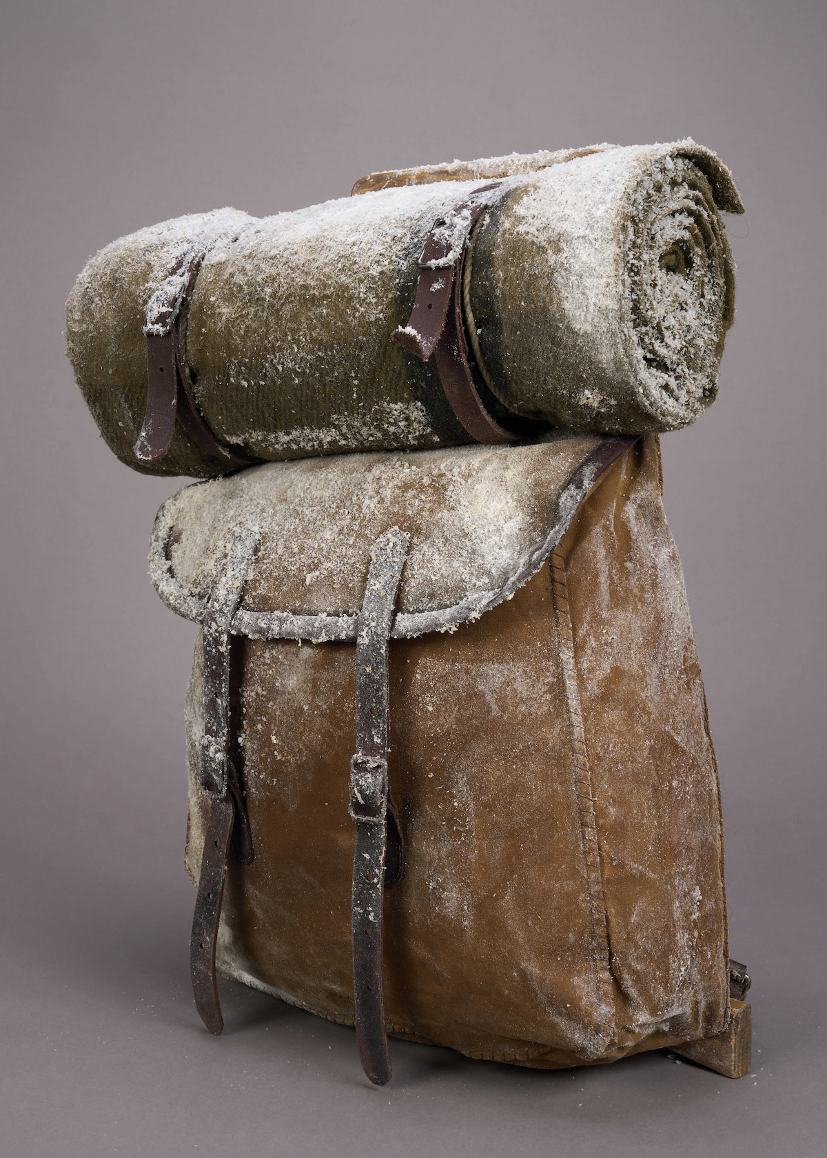 Frost-covered vintage brown backpack with rolled blanket strapped on top, set against a plain gray background, evoking a cold, rugged and adventurous atmosphere.