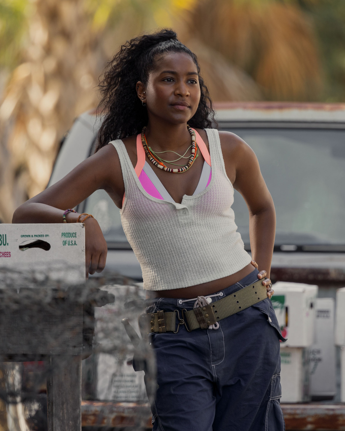 Carlacia Grant as Cleo, wearing layered tank tops and cargo pants leans against a vehicle outdoors, with palm trees and crates in the background, suggesting a warm, casual, and possibly coastal setting.