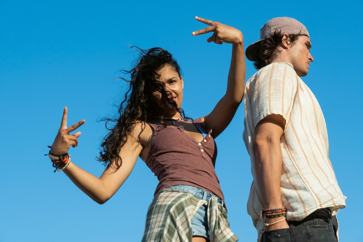 Two young people outdoors, one woman playfully making peace signs and smiling, the other man standing back to back with her, both under a clear blue sky in casual summer clothing.