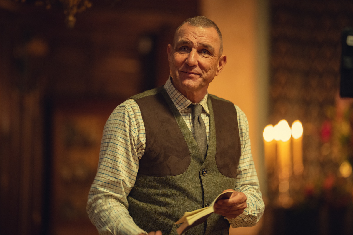 Middle-aged man in a checkered shirt and vest holding a book, standing indoors with warm lighting, blurred background features candles and wooden decor, creating a cozy, intimate atmosphere.