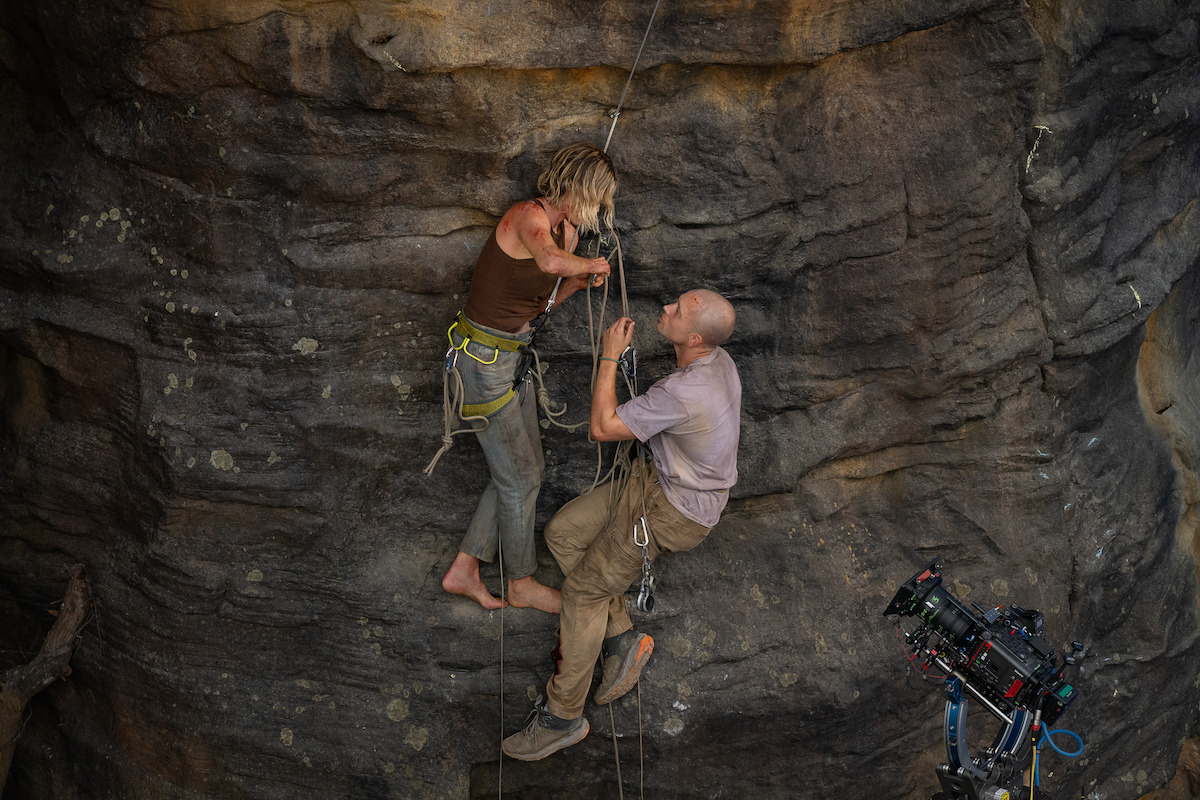 Two people rock climbing on a steep cliff face while a professional film camera captures the scene, indicating a movie or documentary production set in a rugged, outdoor environment.