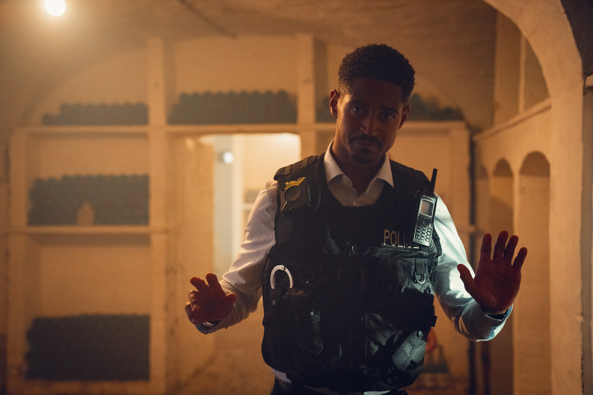 A police officer with hands raised stands in a dim, arched wine cellar, wearing a vest and radio. The environment is tense and atmospheric, with rows of bottles on shelves and dramatic, moody lighting.
