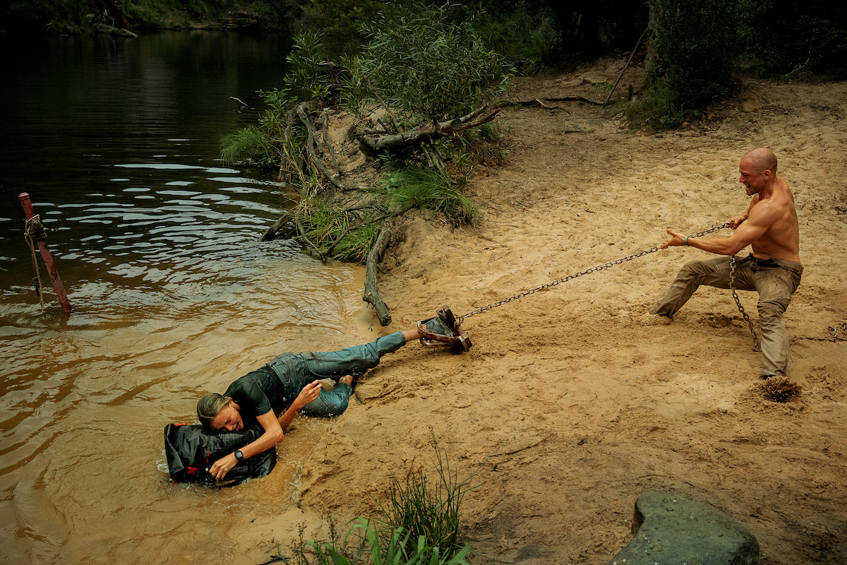 Shirtless man on sandy riverbank pulling another person with a chain from the water, surrounded by trees and bushland, intense survival or rescue scene.