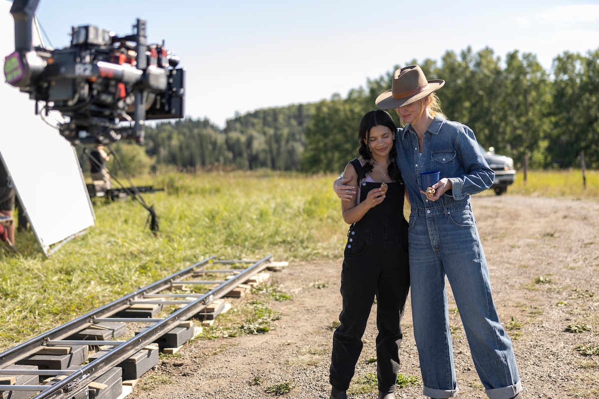 Two women stand together outdoors on a film set, looking at a phone, with camera equipment and a grassy landscape in the background.