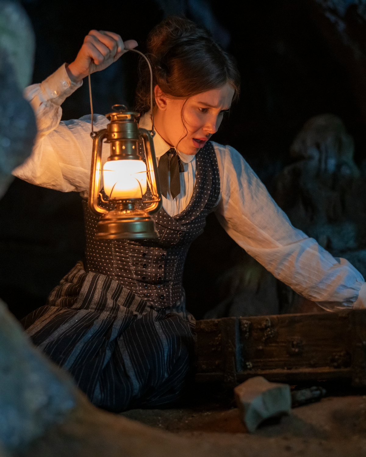 A young woman in vintage clothing kneels in a dark cave, holding a lit lantern while looking into an open wooden chest, surrounded by rocks, suggesting exploration or discovery in a mysterious underground setting.