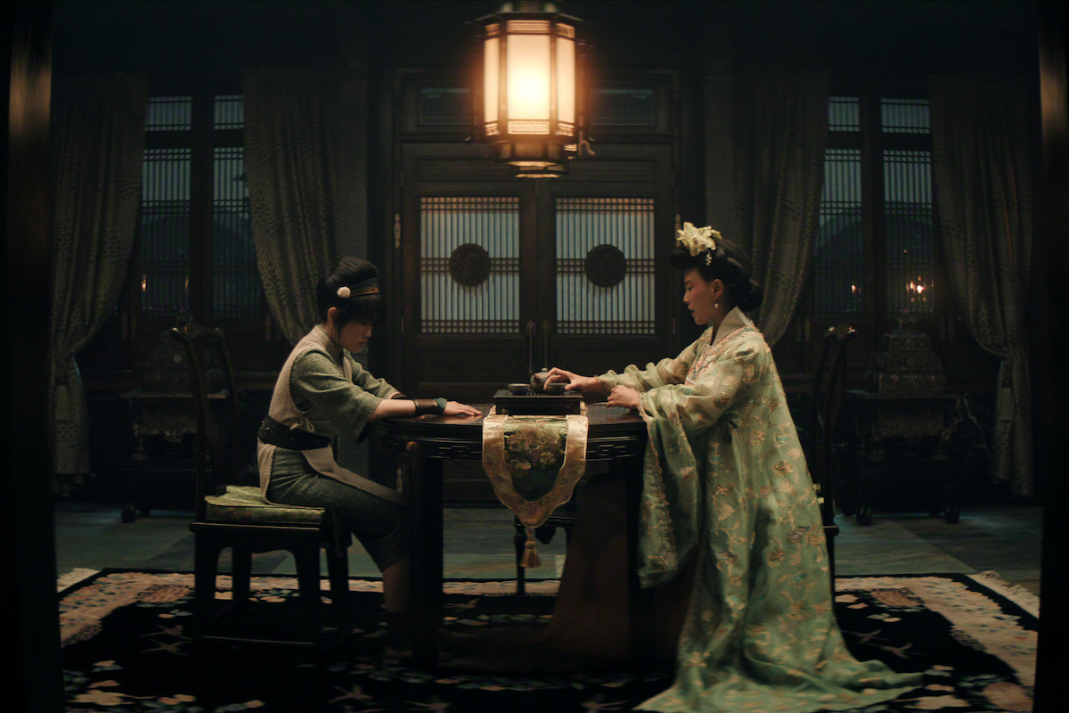 Miyako as Toph and Crystal Yu as Lady Beifong sit across from each other at an ornate table in an elegant, dimly lit room with elaborate wooden doors and a large hanging lantern above.