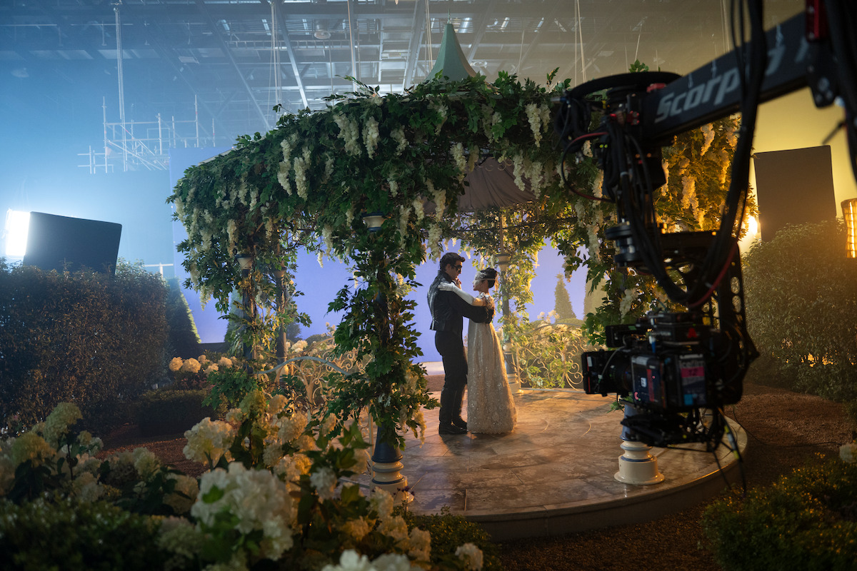 A couple dances under a flower-covered gazebo on a film set surrounded by greenery, with studio lighting and a large camera capturing the scene.