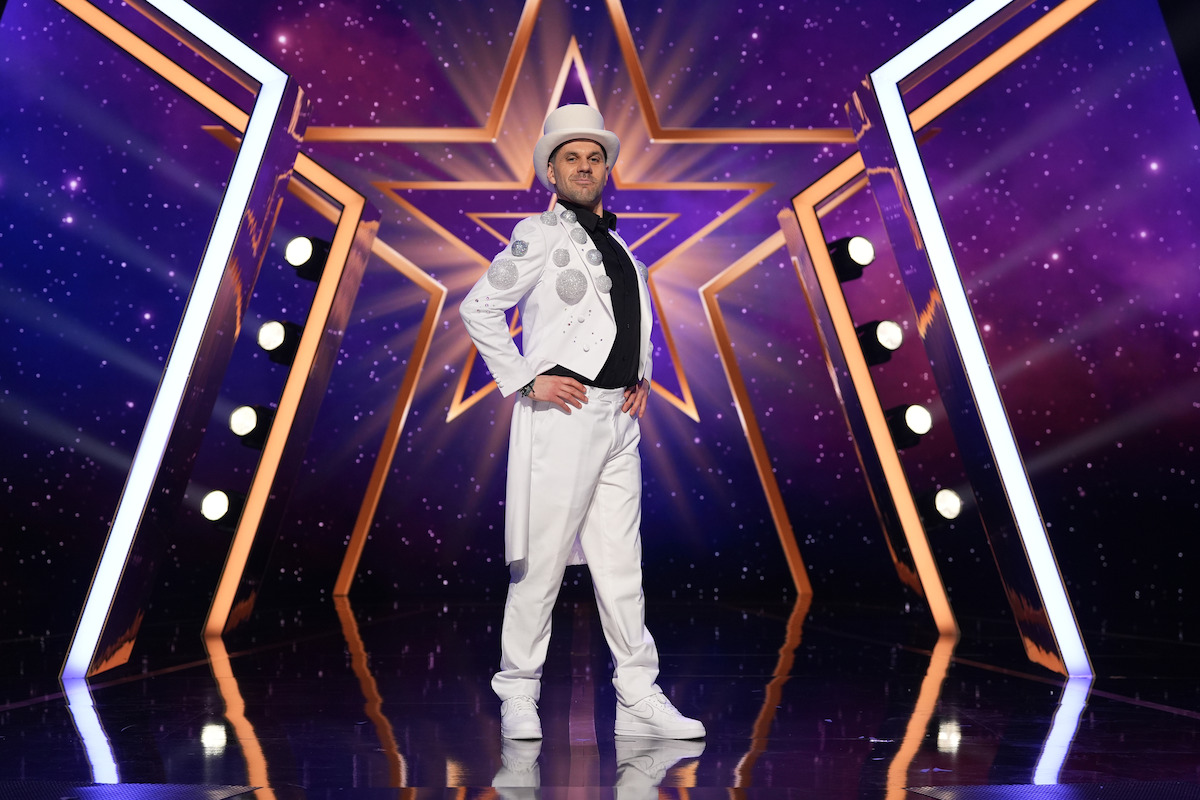 Luca Cutrupi in a white costume and hat poses confidently on a shiny stage with star-themed purple and gold lighting and geometric props in the background, suggesting a performance or talent show setting.