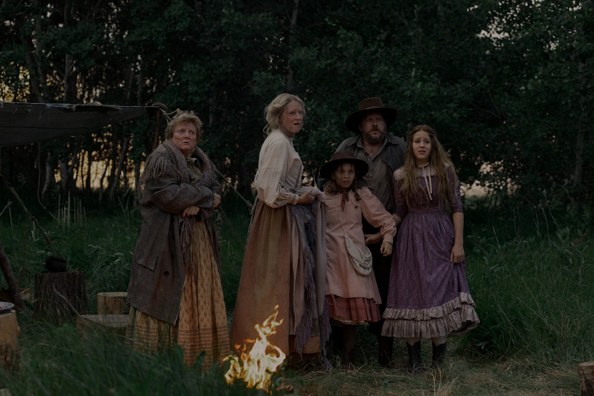 Five people in old-fashioned clothing stand together outside in tall grass near a campfire and tent, surrounded by trees at dusk, suggesting a historical, rustic setting.
