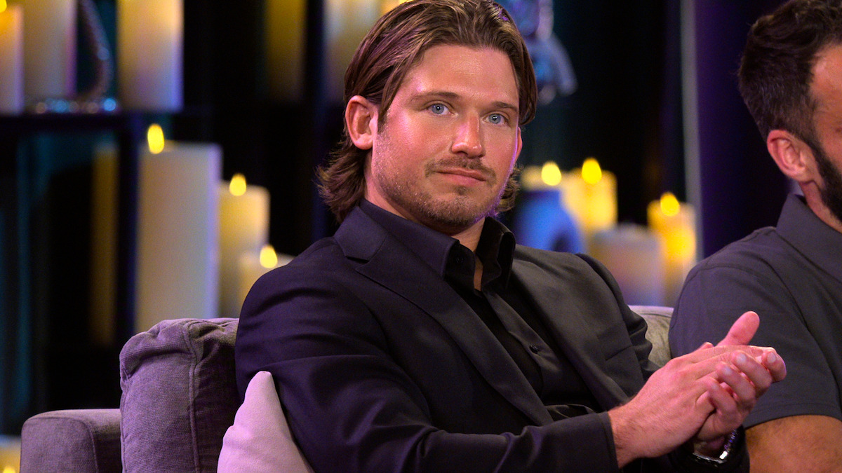 Man in a black suit sitting on a gray couch, clapping his hands, with many lit candles in the background creating a warm, intimate atmosphere.