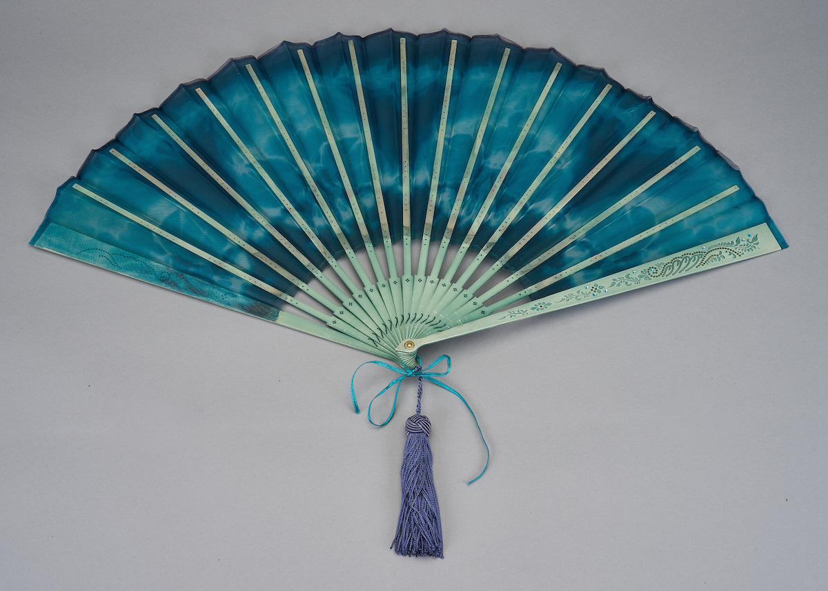 A decorative hand fan with blue and green hues, light wooden ribs, a blue tassel, and ribbon, displayed open against a plain gray background, evoking elegance and a tranquil, refined mood.