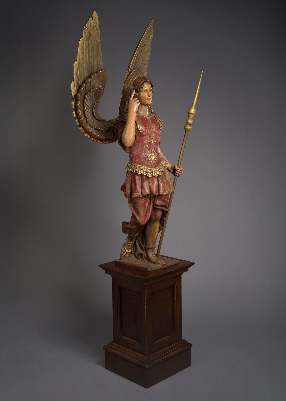A statue of a winged figure in armor holding a spear, set on a pedestal in a dimly lit, plain indoor environment. The mood is solemn and mysterious, with muted colors emphasizing the figure's imposing presence.