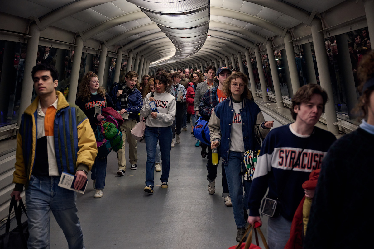 A group of college students wearing Syracuse sweatshirts and jackets walk through a tunnel with curved ceiling and glass walls, carrying bags and backpacks, appearing to travel or return from an event.